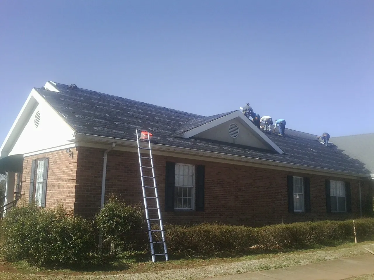 Crew tearing shingles off a brick ranch roof with a ladder set — Metal Roof Repair work by our crew in Blue Mounds