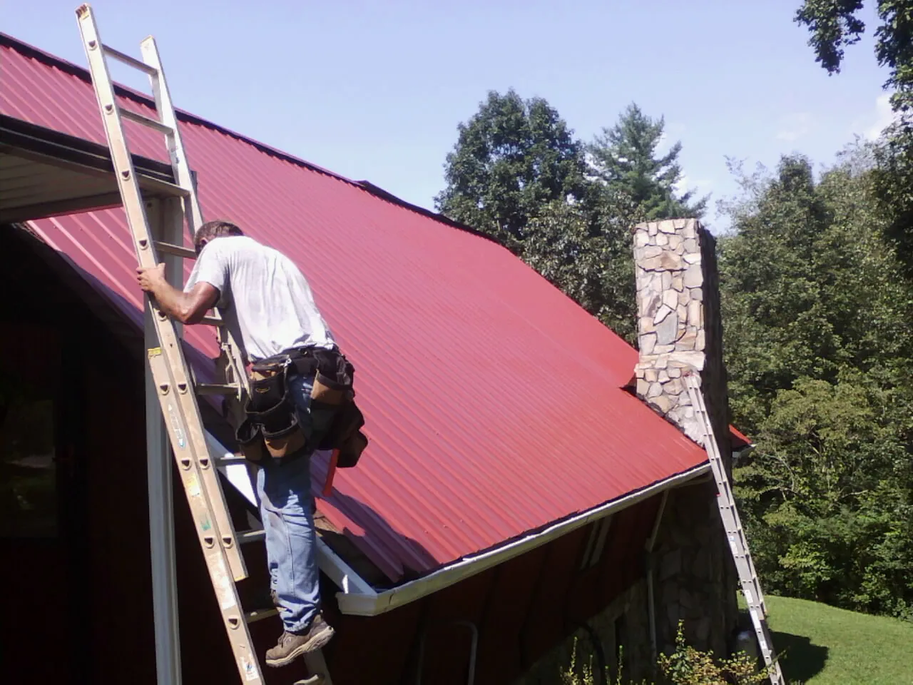 Roofer on a ladder beside a red metal roof and stone chimney — Soffit & Fascia Repair by Roofing Brothers in Blue Mounds